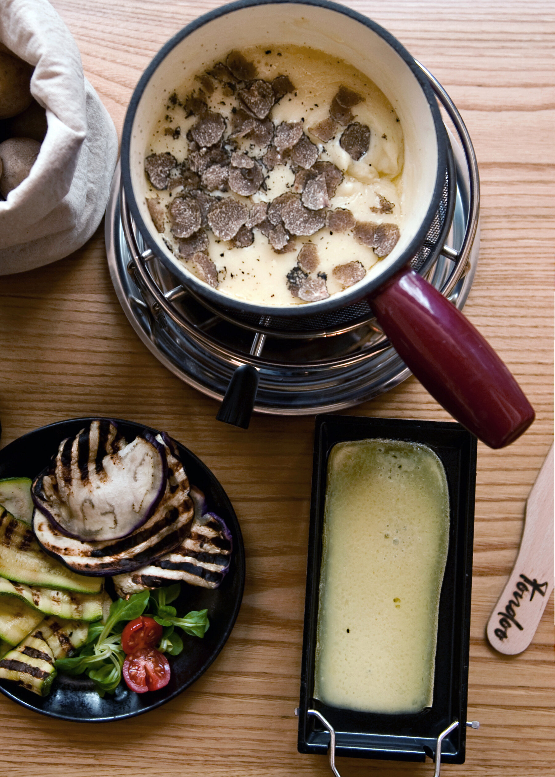 A wooden table is laden will small dishes. There is a small plate of grilled eggplant and zucchini; a plate with a wedge of cheese; and a fondue pot with melted cheese and shaved truffle. 