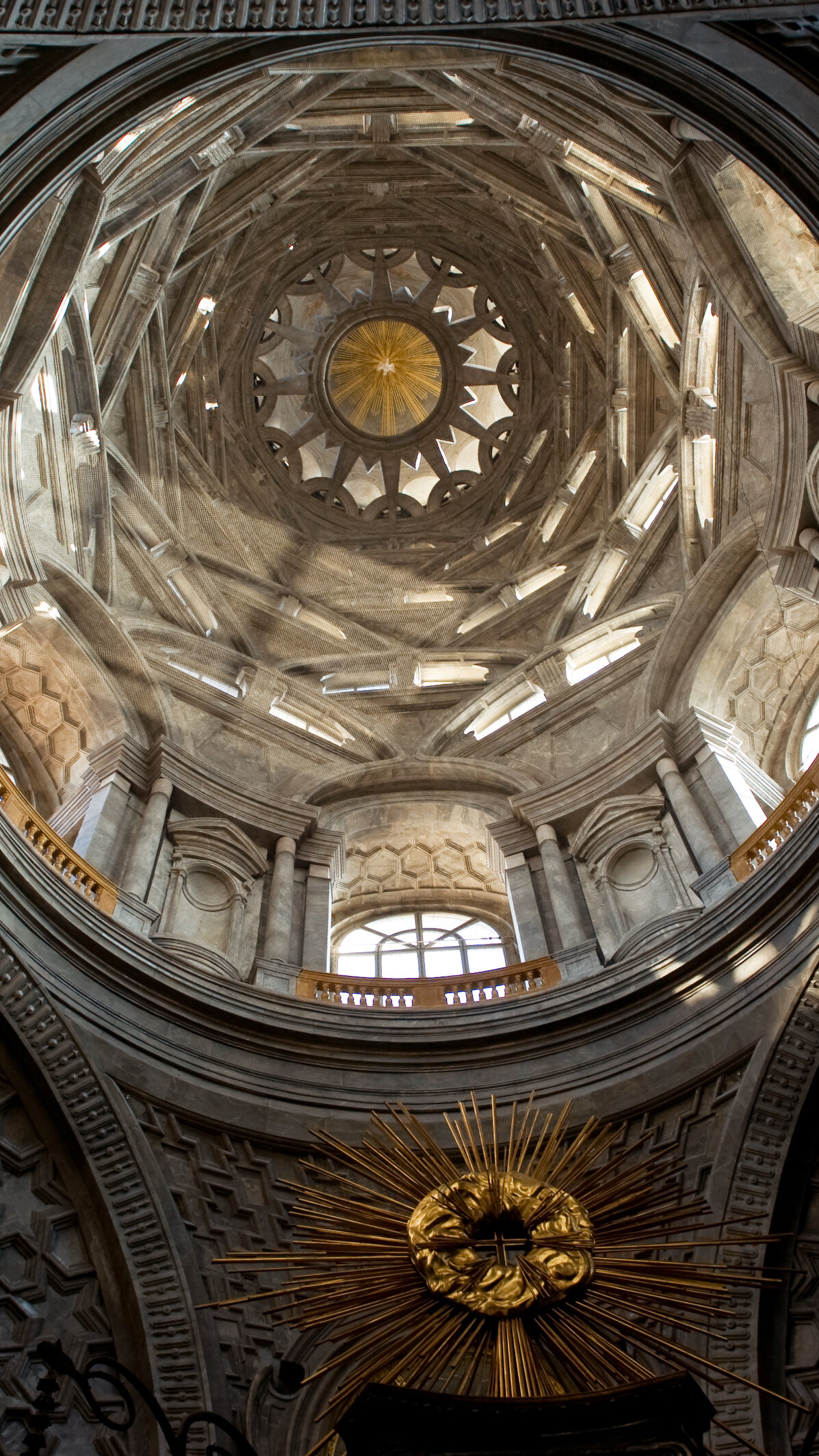 A view looking up at the interior of an ornately decorated dome in a grand building. Sunlight is streaming through windows at the top of the dome. 