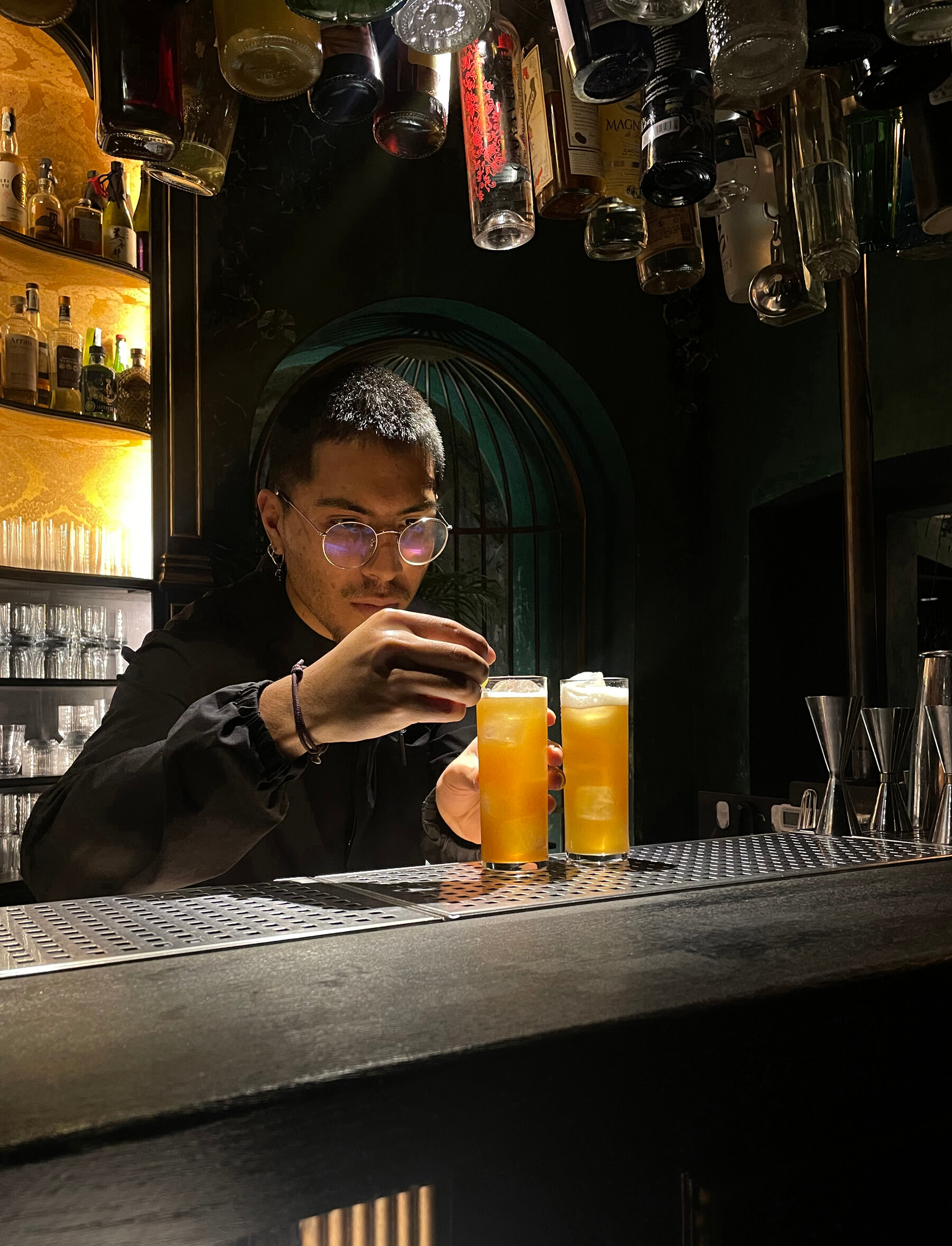 A person with round spectacles makes two drinks in highball glasses from behind a black bar top. Liquor bottles hang above the person. 