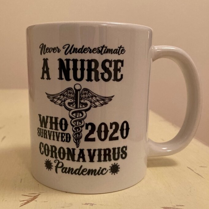A white mug reading "Never Underestimate A Nurse Who Survived 2020 Coronavirus Pandemic" sits on a cream-colored countertop.