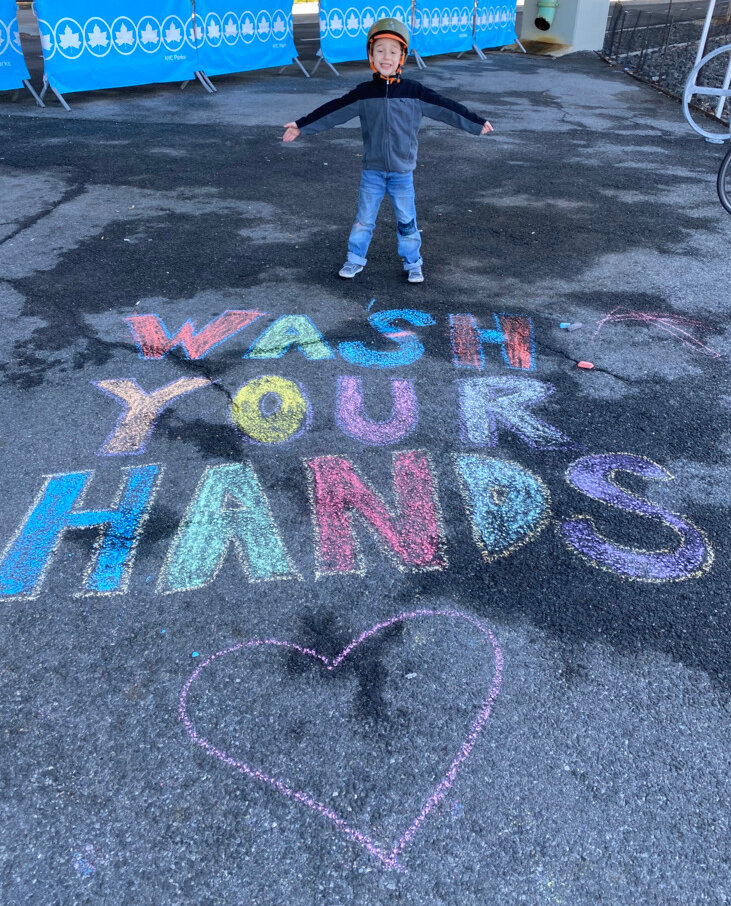 A boy standing in behind words written in chalk on the pavement. The message reads “Wash Your Hands” with a heart underneath.