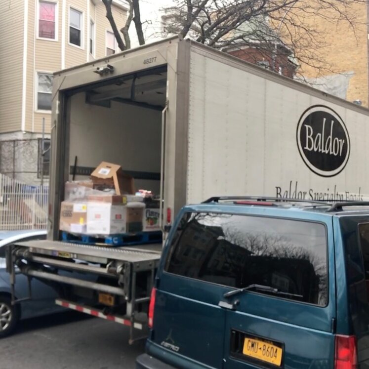A box truck parked in the middle of a street with its back door open. A logo on the side reads “Baldor.” There are packages sitting inside the truck.