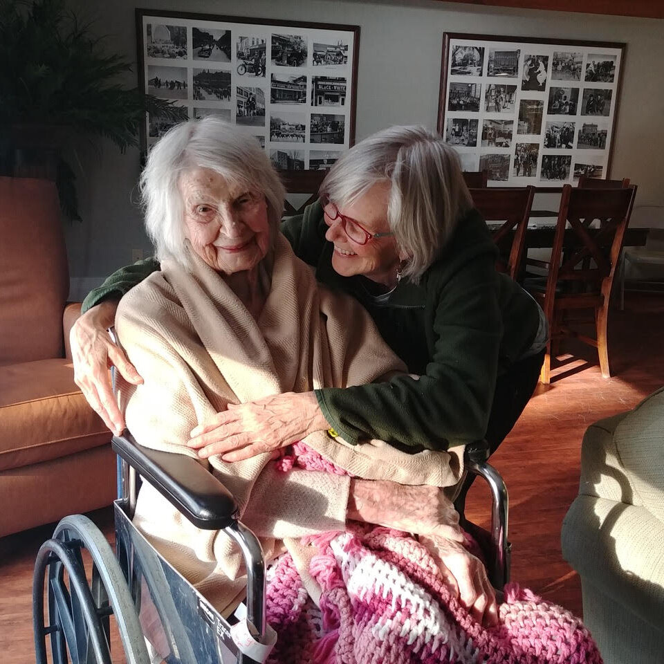 A woman hugging an older woman who is sitting in a wheelchair in a residential setting. Sunlight is beaming onto them.