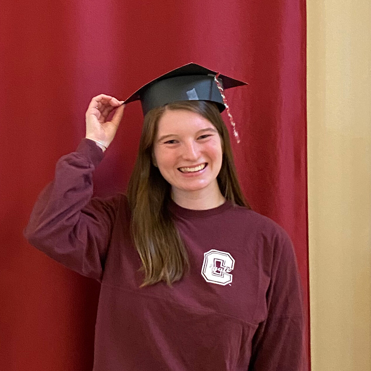 A young woman standing in front of a red backdrop with a homemade graduation cap on her head. Her maroon sweatshirt has the letter “C” on it.