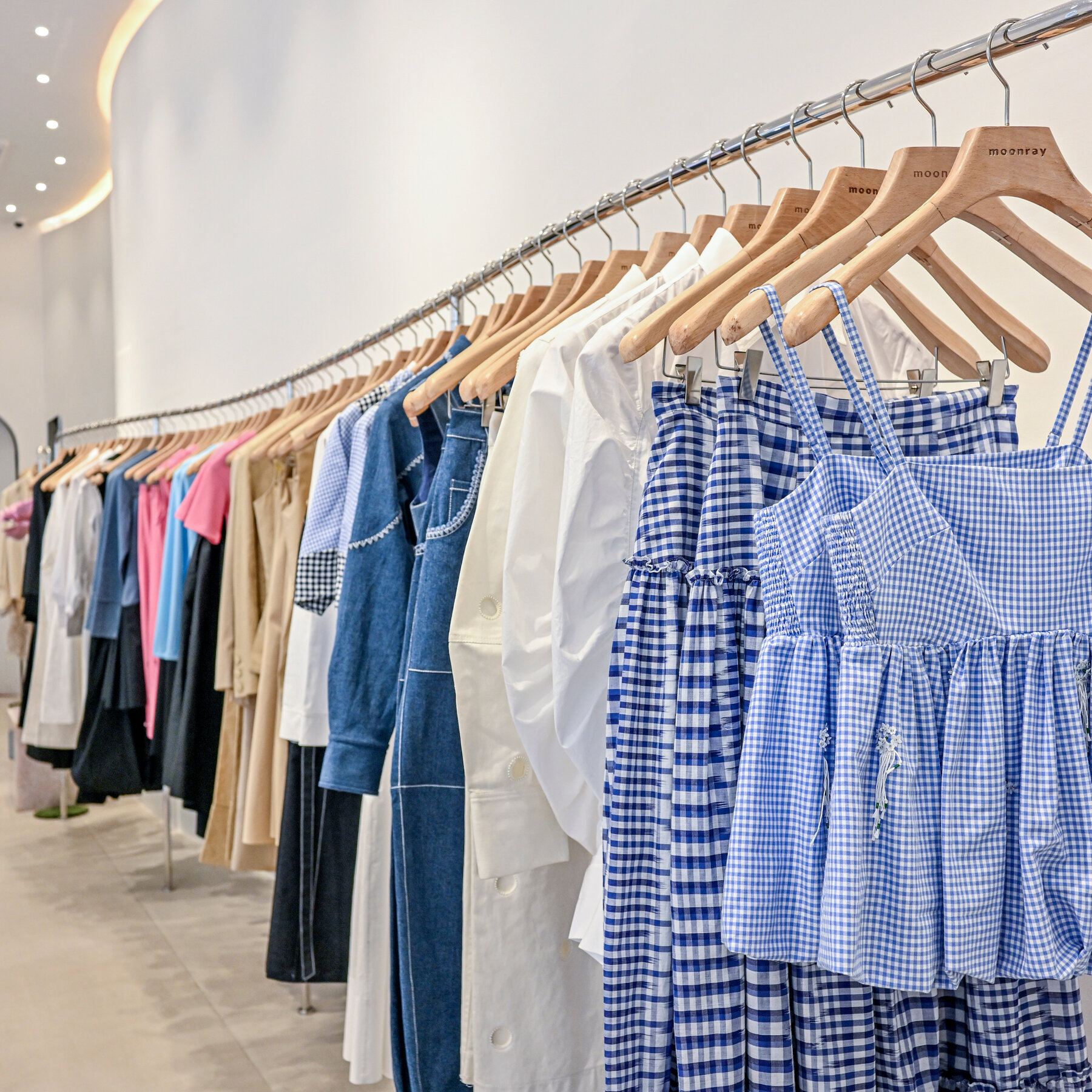 A brightly lit store with white walls displays a long rack of clothing, including gingham skirts and tops, white shirts and denim apparel.