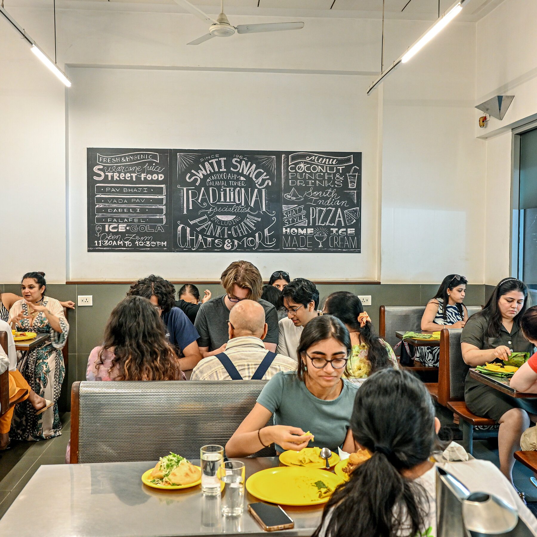 People eat from bright-yellow plates in a sparsely decorated restaurant with diner-like seating and metallic table tops. A chalkboard on the wall lists menu items and says the name of the restaurant, "Swati Snacks."