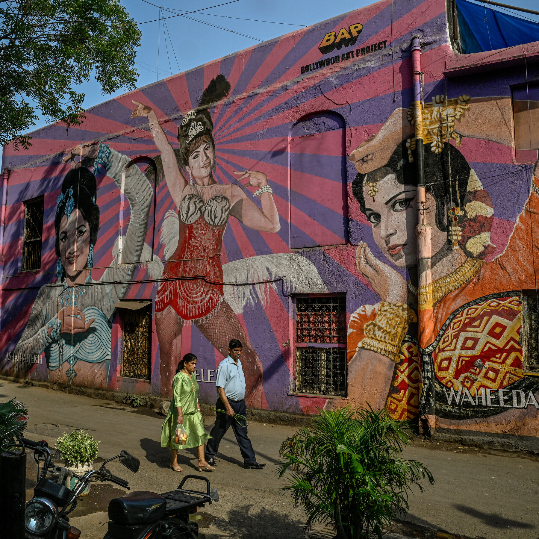 Two people walk past a vibrant street mural of three dancing women dressed in ornate Indian dress.