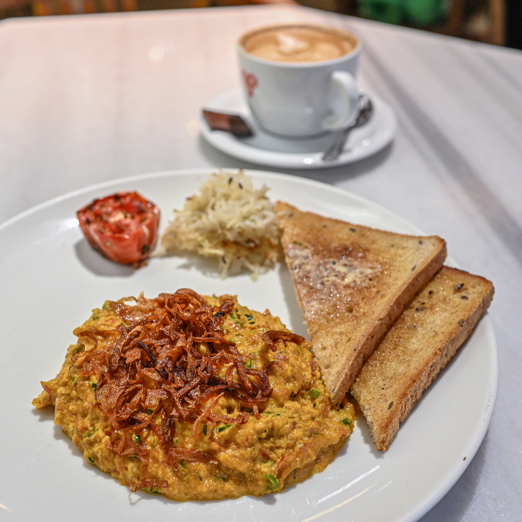 A plate of scrambled eggs, buttered toast and half a roasted tomato rests on a table. A cup of coffee with steamed milk is visible in the background on the same table. 