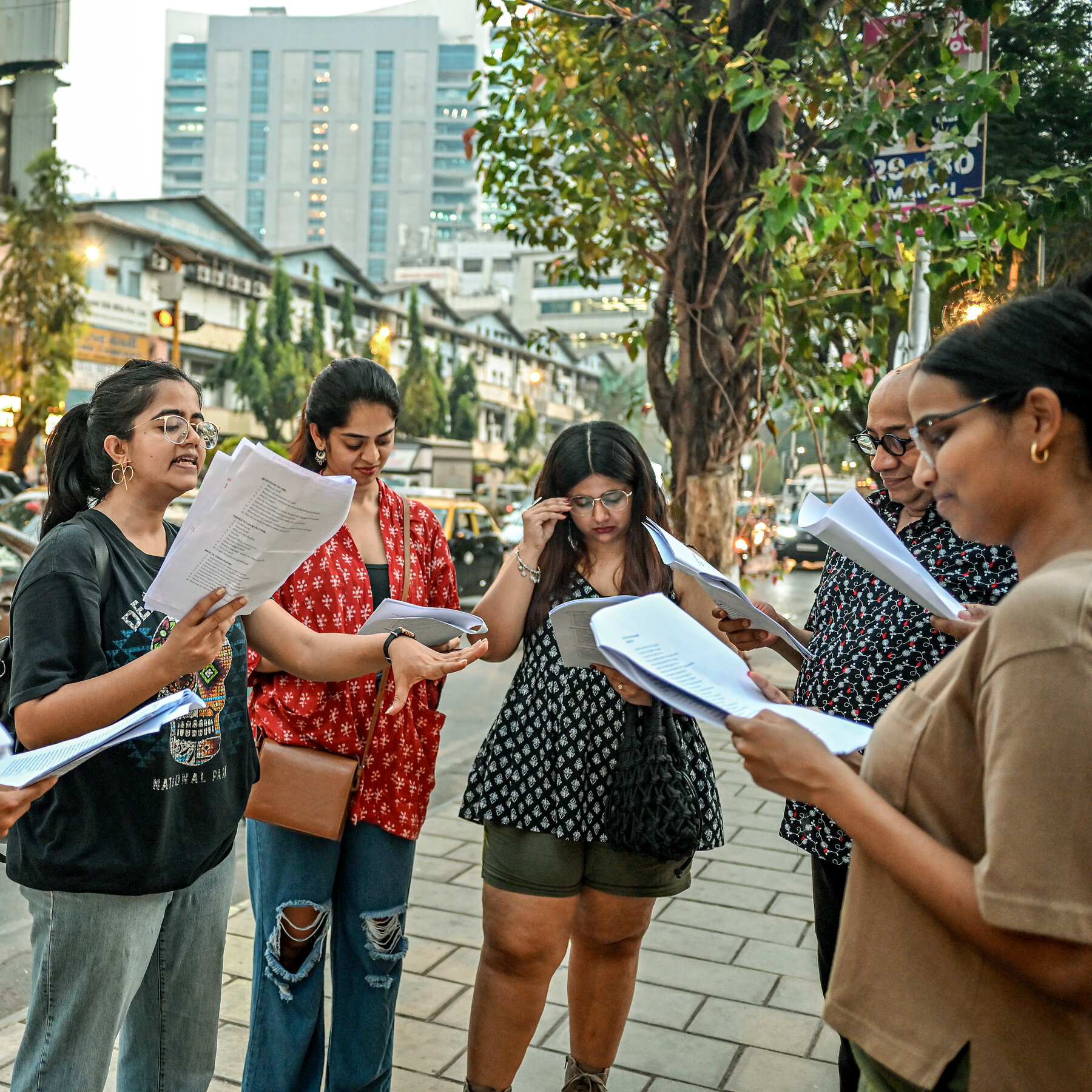 People stand in a semi-circle on a leafy street reading from a stapled paper booklet. 