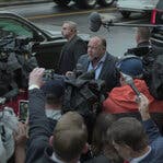 Alex Jones stands amid a group of reporters with microphones near a black SUV on the street.