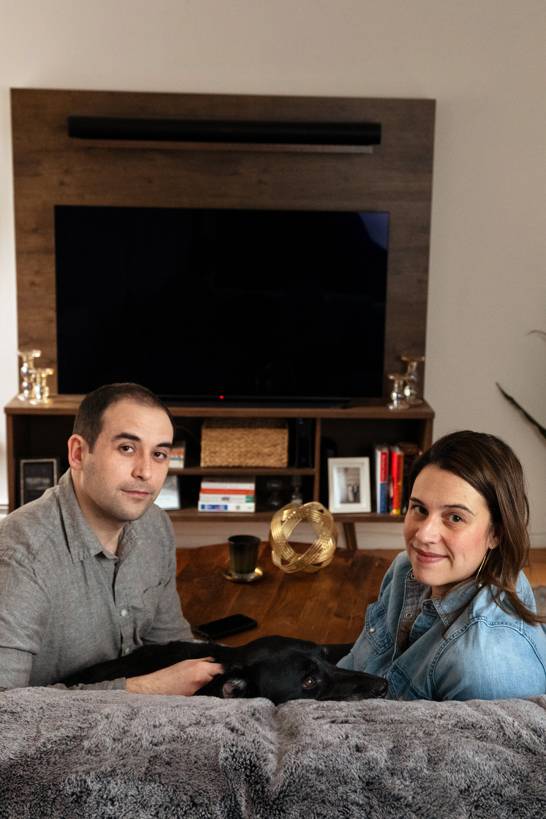 A young couple sit on the sofa of a living room and look at the camera, with a black dog between them.