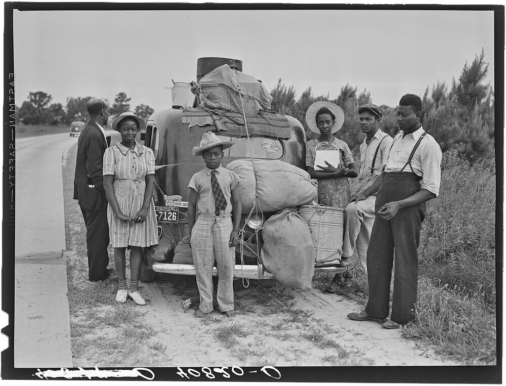 In a black and white image from 1940, a group of people of varying ages stand on the side of a road next to a car loaded down with bags and belongings.