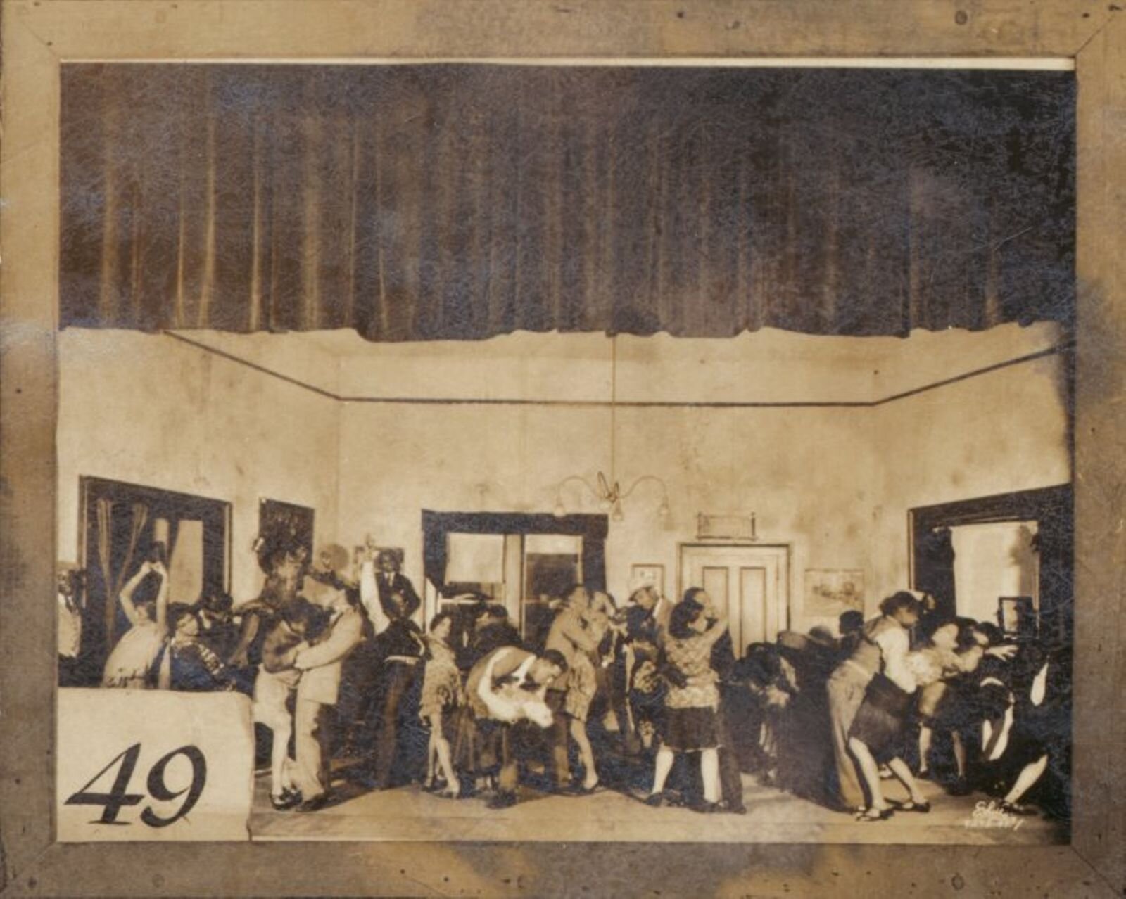 A sepia-colored photo shows a group of actors dancing in a party scene on a stage.