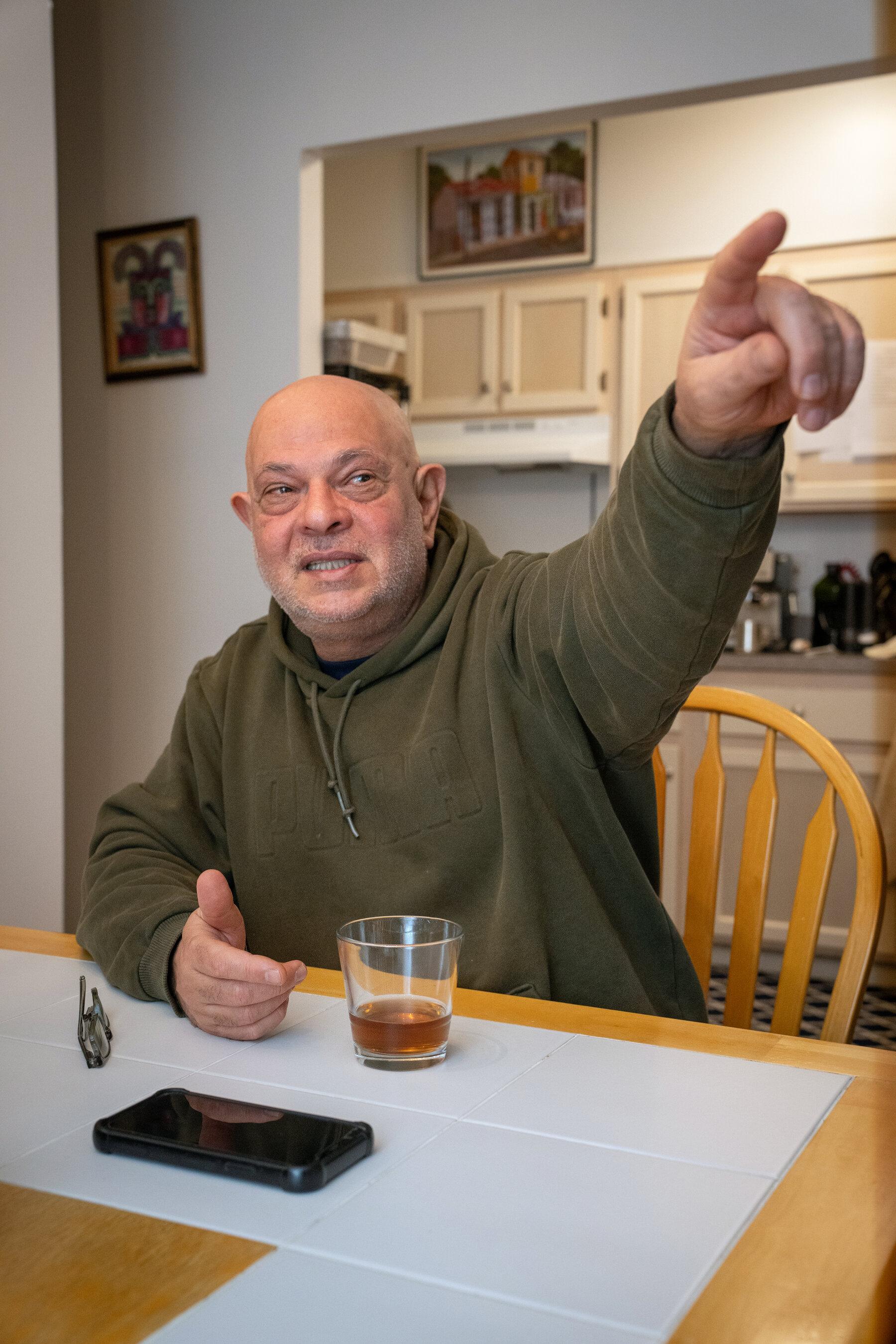 A man in an olive green sweatshirt sits at a kitchen table and points.