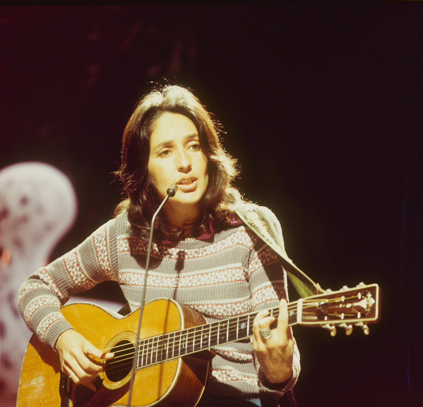 Joan Baez holds a guitar and sings into a microphone.