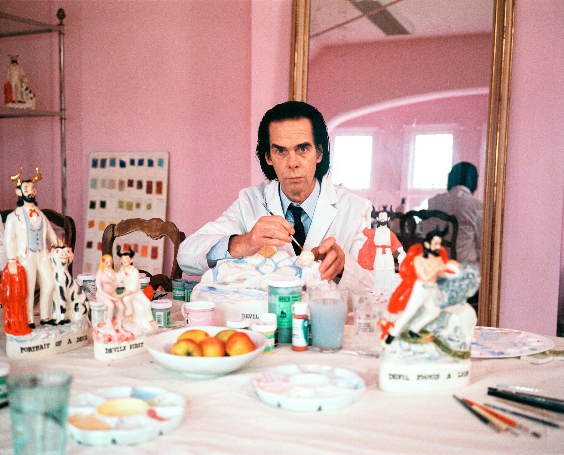 Nick Cave, wearing a shirt, tie and white jacket and sitting in a pink room in front of a tall mirror, holds a paint brush above a porcelain figure. In front of him, on the table, are paint palates, a bowl of fruit and various sculptures.