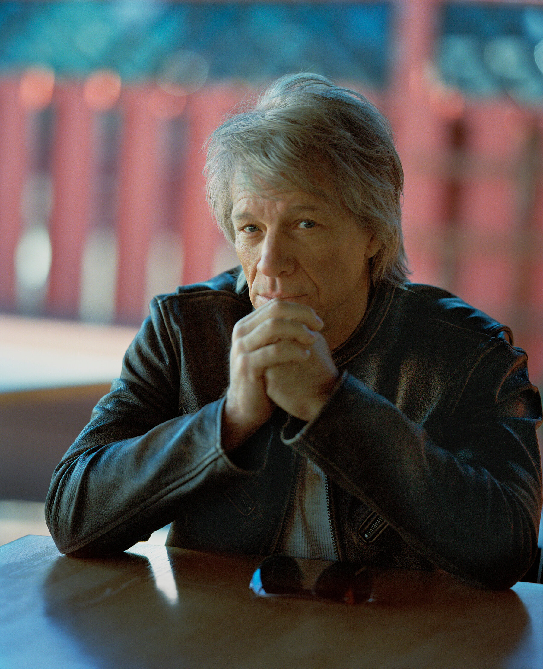 Bon Jovi, with gray hair pushed to one side, wears a leather jacket and leans his elbows on a wooden table and looks into the camera.