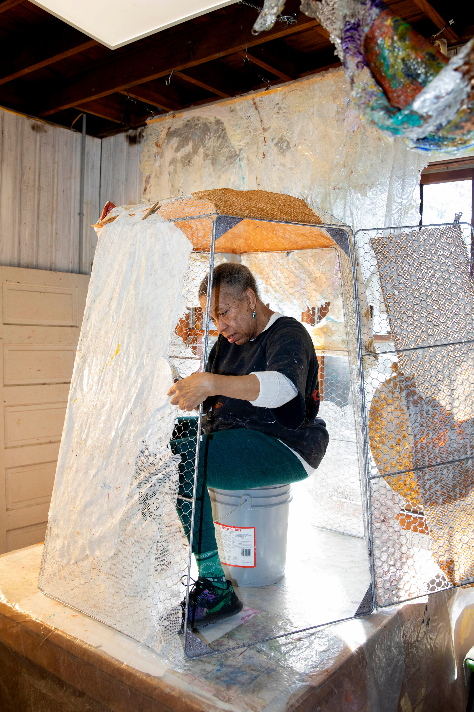 Suzanne Jackson sits on a bucket assembling a sculptural work involving paper or plastic and wire mesh.