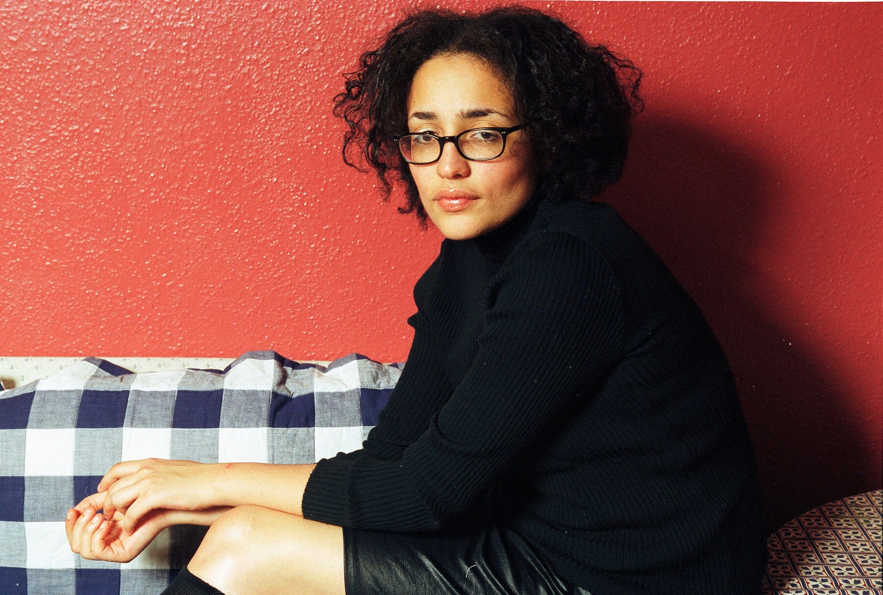 Zadie Smith, wearing a black top and glasses, with her hair parted in the center, sits and looks over her left shoulder towards the camera. The wall behind her is red.