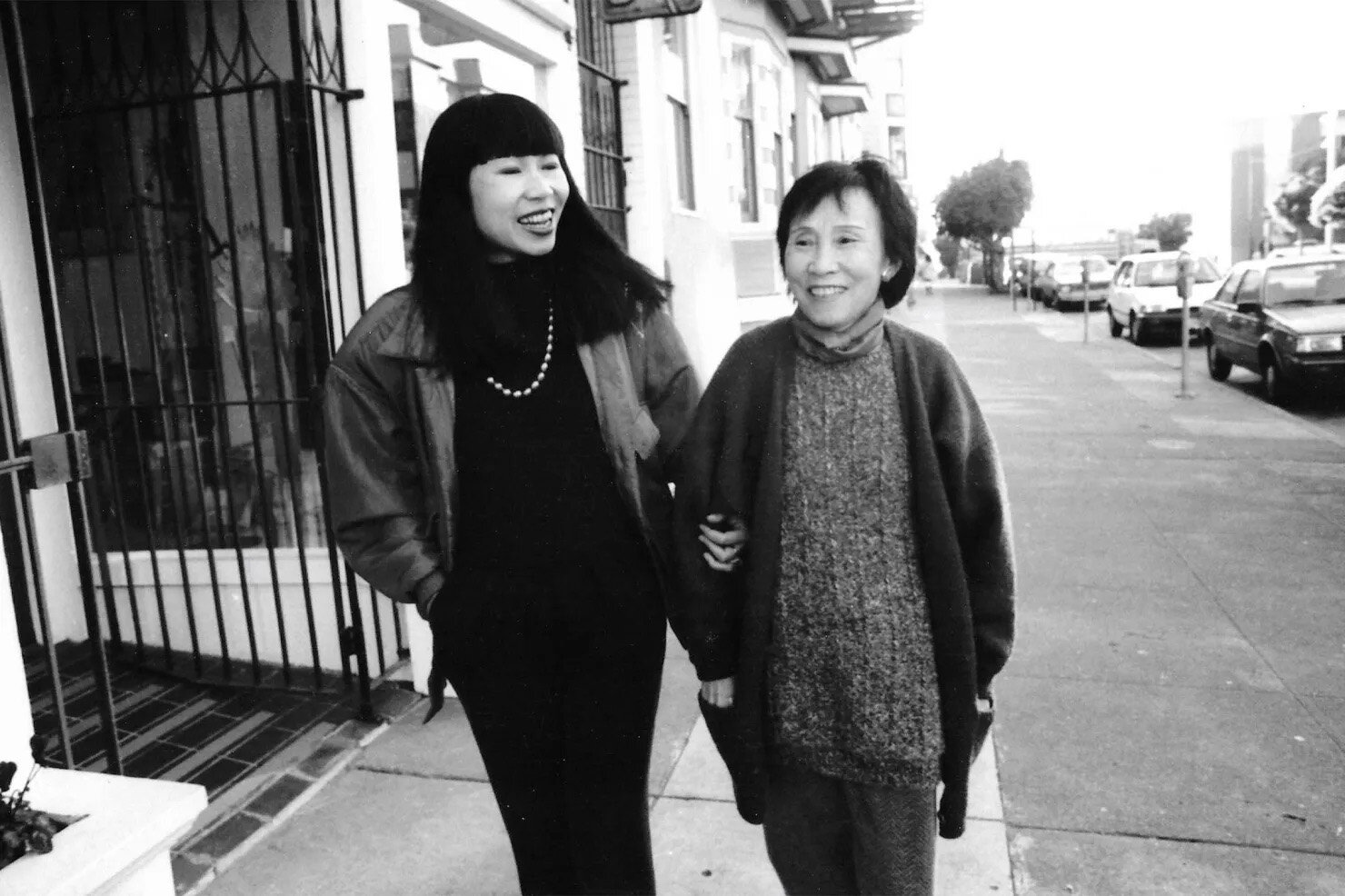 Amy Tan holds Daisy Tan's right elbow with her left hand. They are walking down the a sidewalk and smiling.