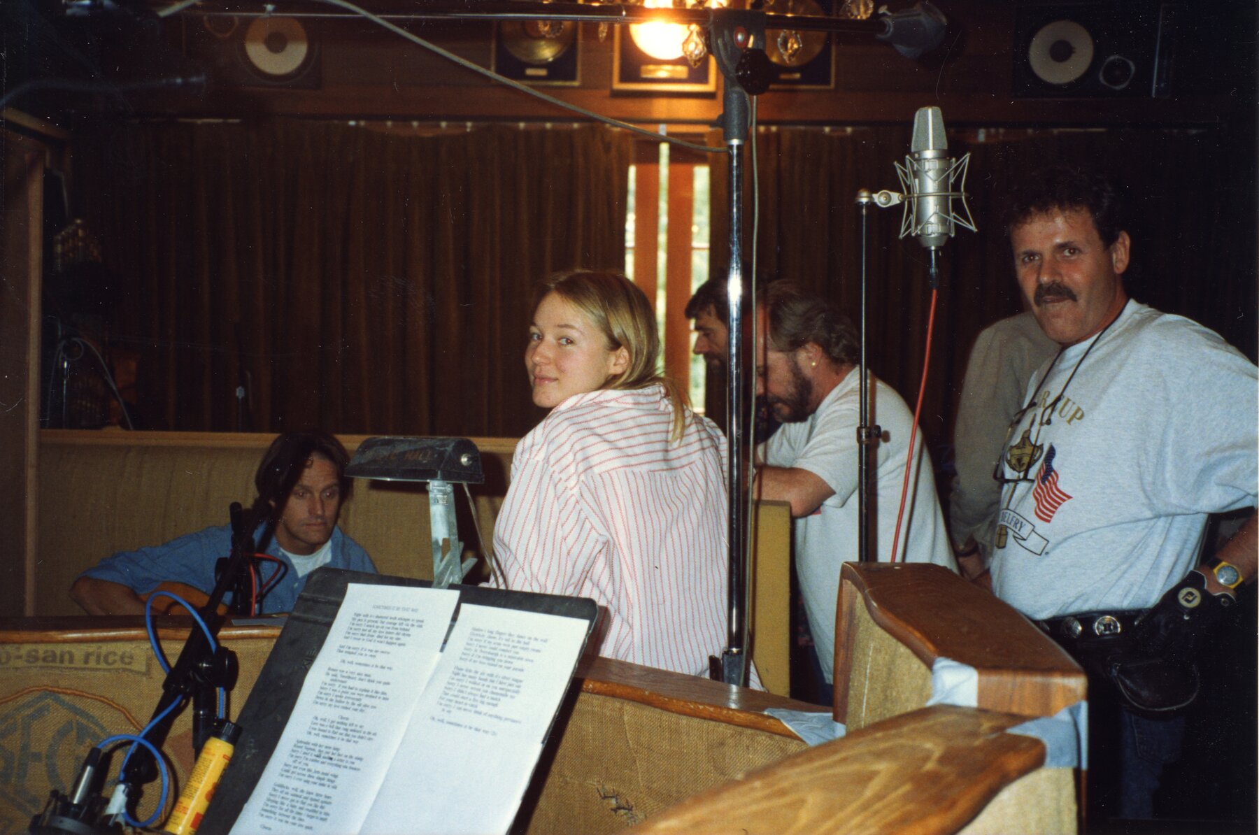 Jewel, surrounded by people in a recording studio, wearing a white and orange striped shirt, looks back over her left shoulder.