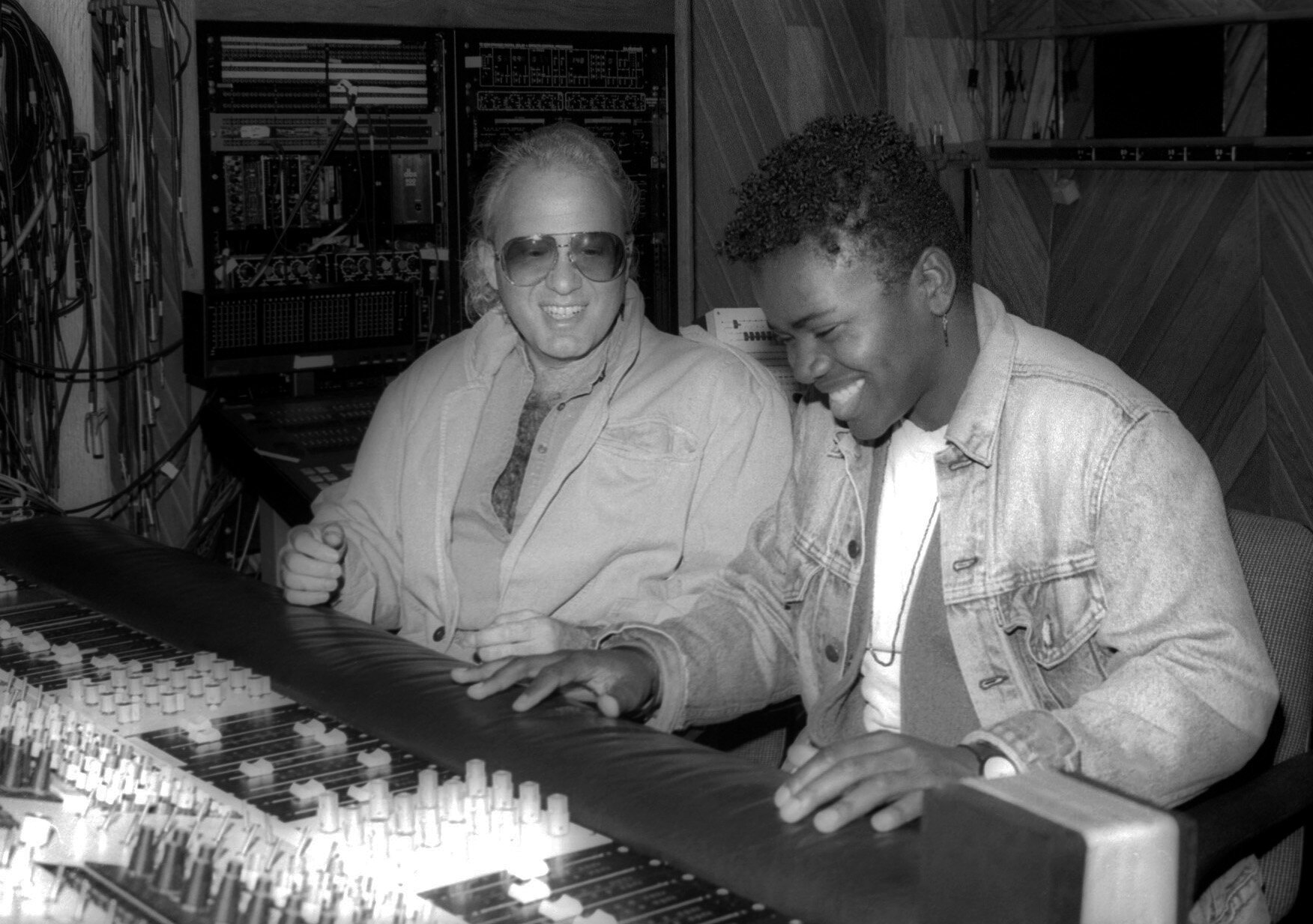 David Kershenbaum, wearing an open shirt and sunglasses, sits next to Tracy Chapman, wearing a jean jacket, in front of a control board in a recording studio.