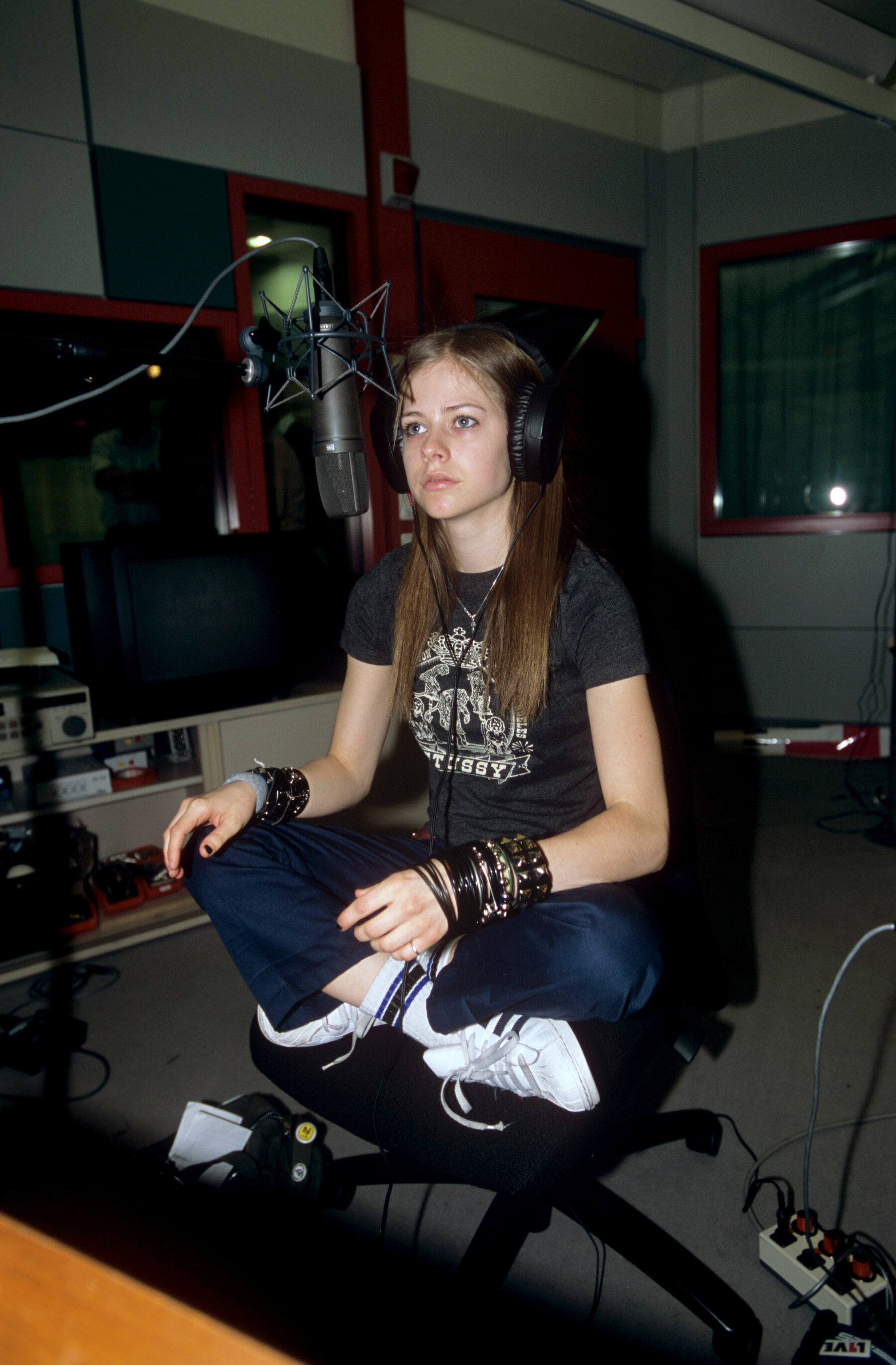 Avril Lavigne sits cross-legged on an office chair wearing headphones with a microphone in front of her face.