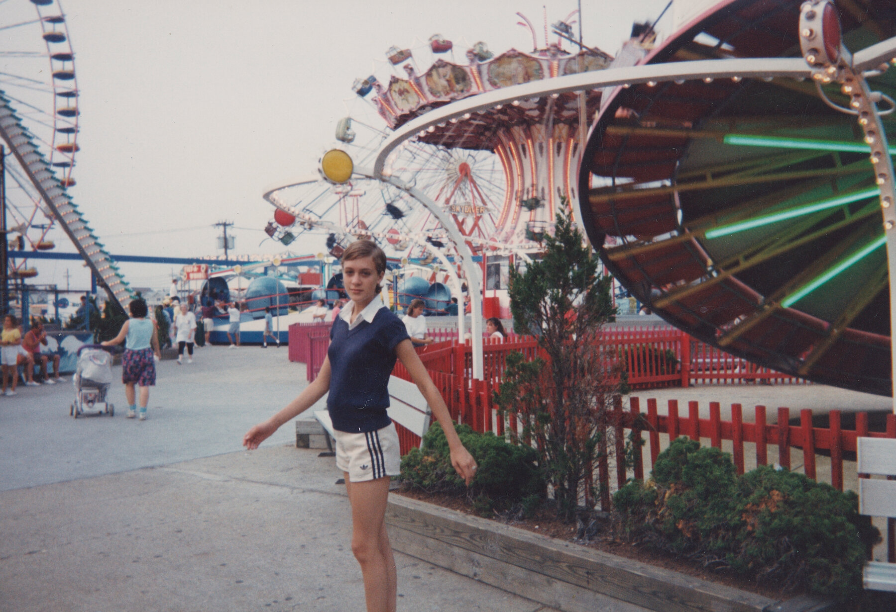 Chloë Sevigny turns to face the camera. Behind her are various theme park attractions, including a ferris wheel and a carousel.