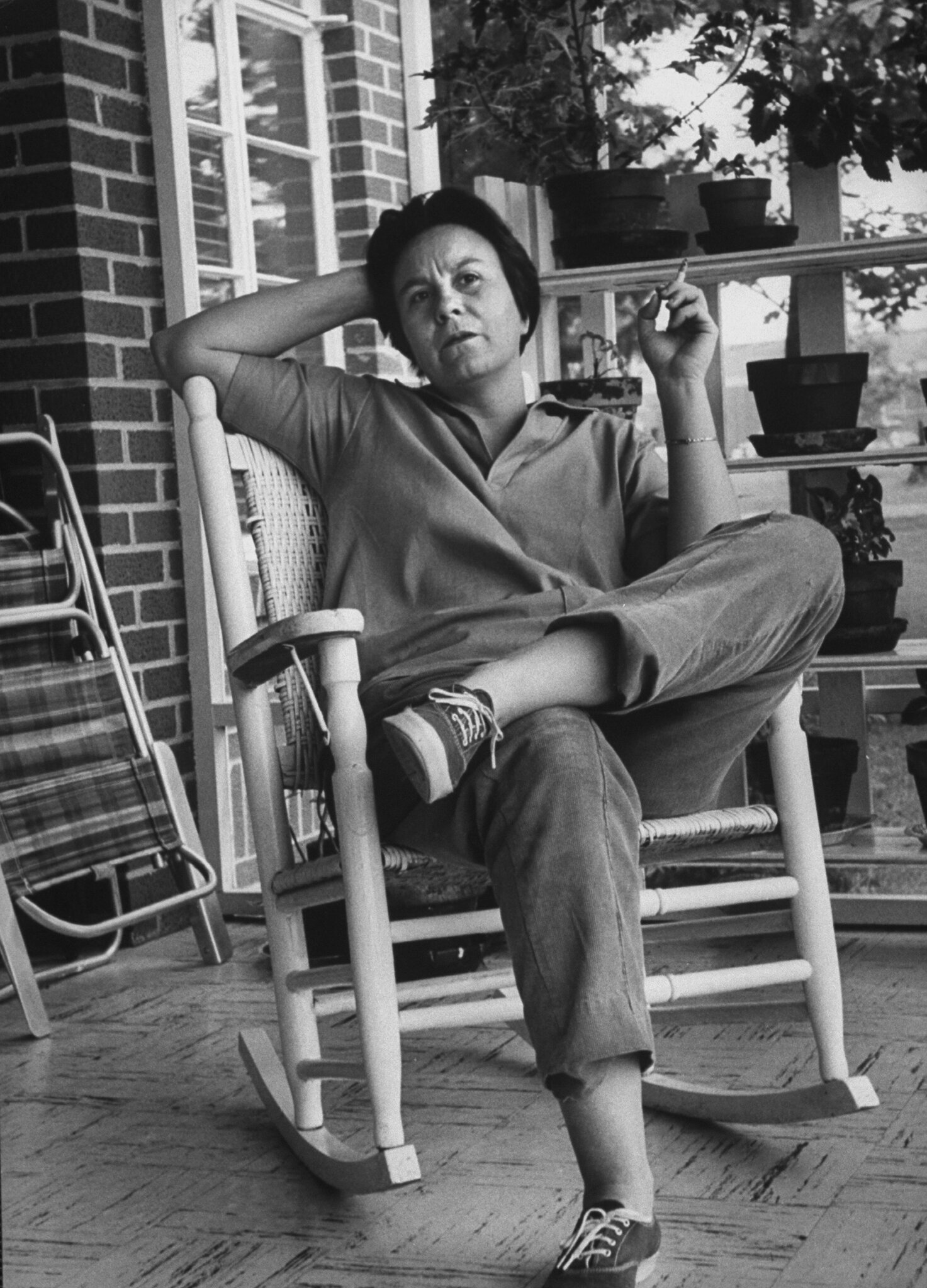 A portrait of Harper Lee sitting on a rocking chair on a porch smoking a cigarette.