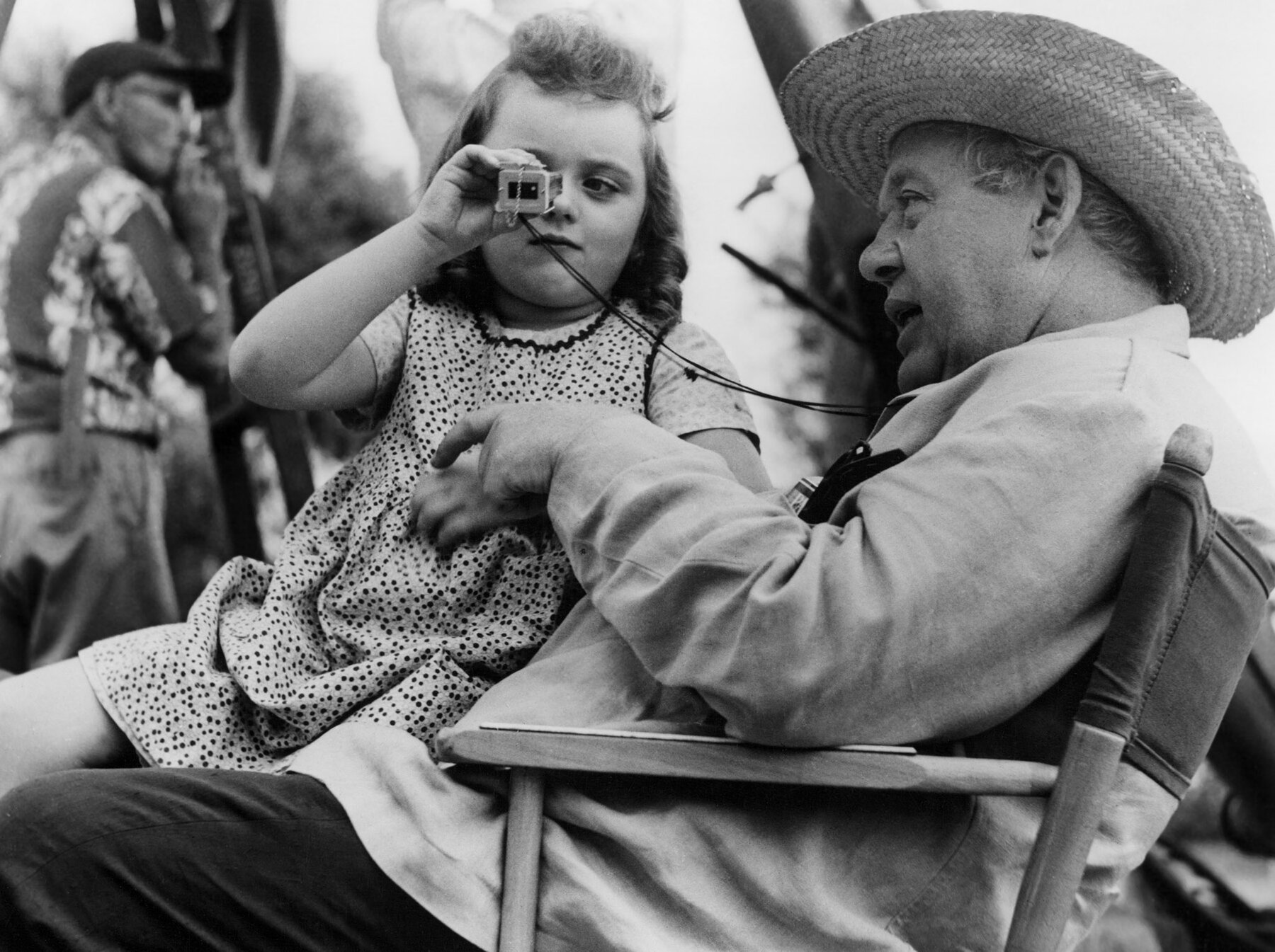 Charles Laughton sits in a director's chair wearing a straw hat with a girl looking through a viewfinder in his lap.