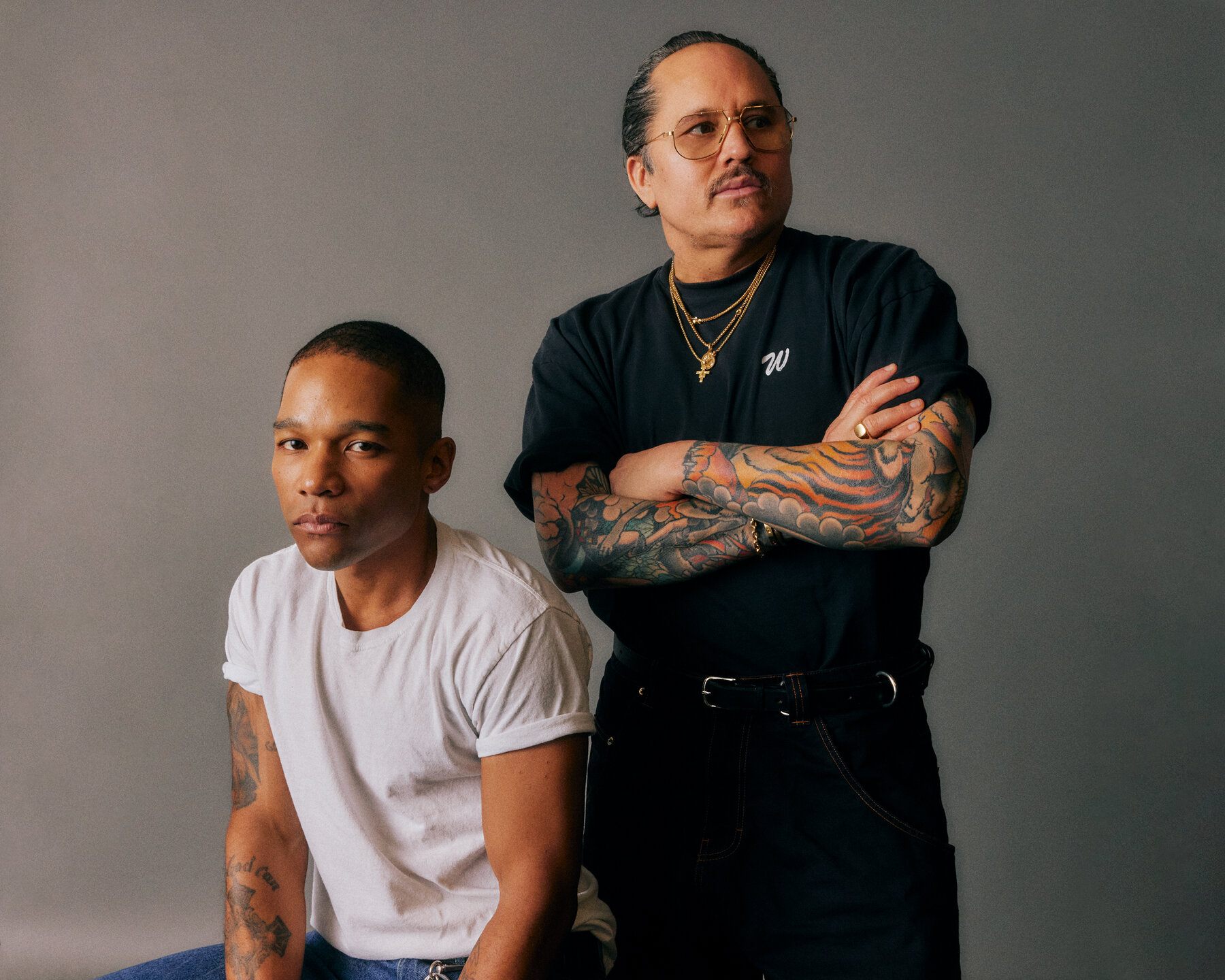 Willy Chavarria, wearing a black T-shirt and necklaces, stands and crosses his arms. Next to him sits Carlos Nazario, wearing a white T-shirt.