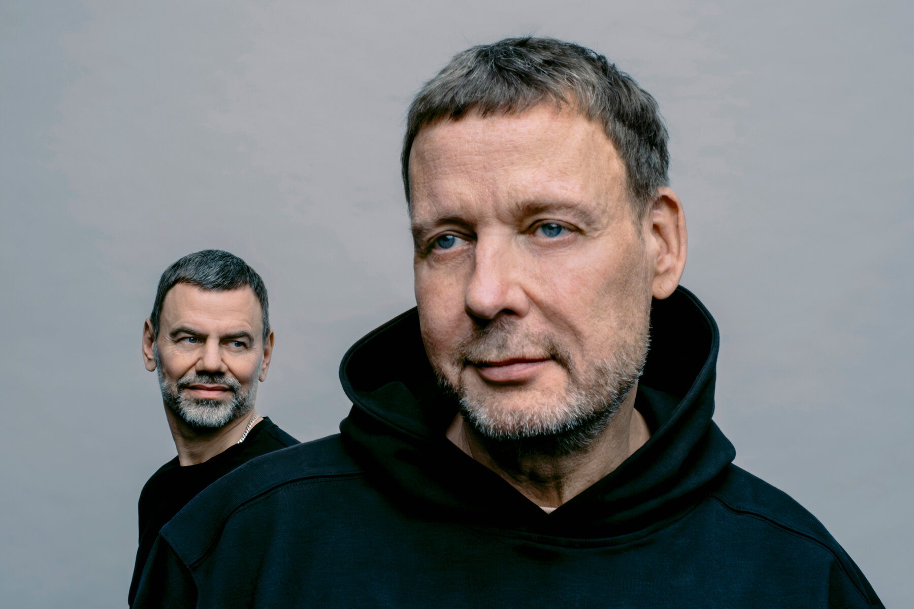 A portrait of Dragset and Elmgreen smiling and standing in front of a gray background. Dragset wears a black T-shirt and Elmgreen wears a black hoodie.