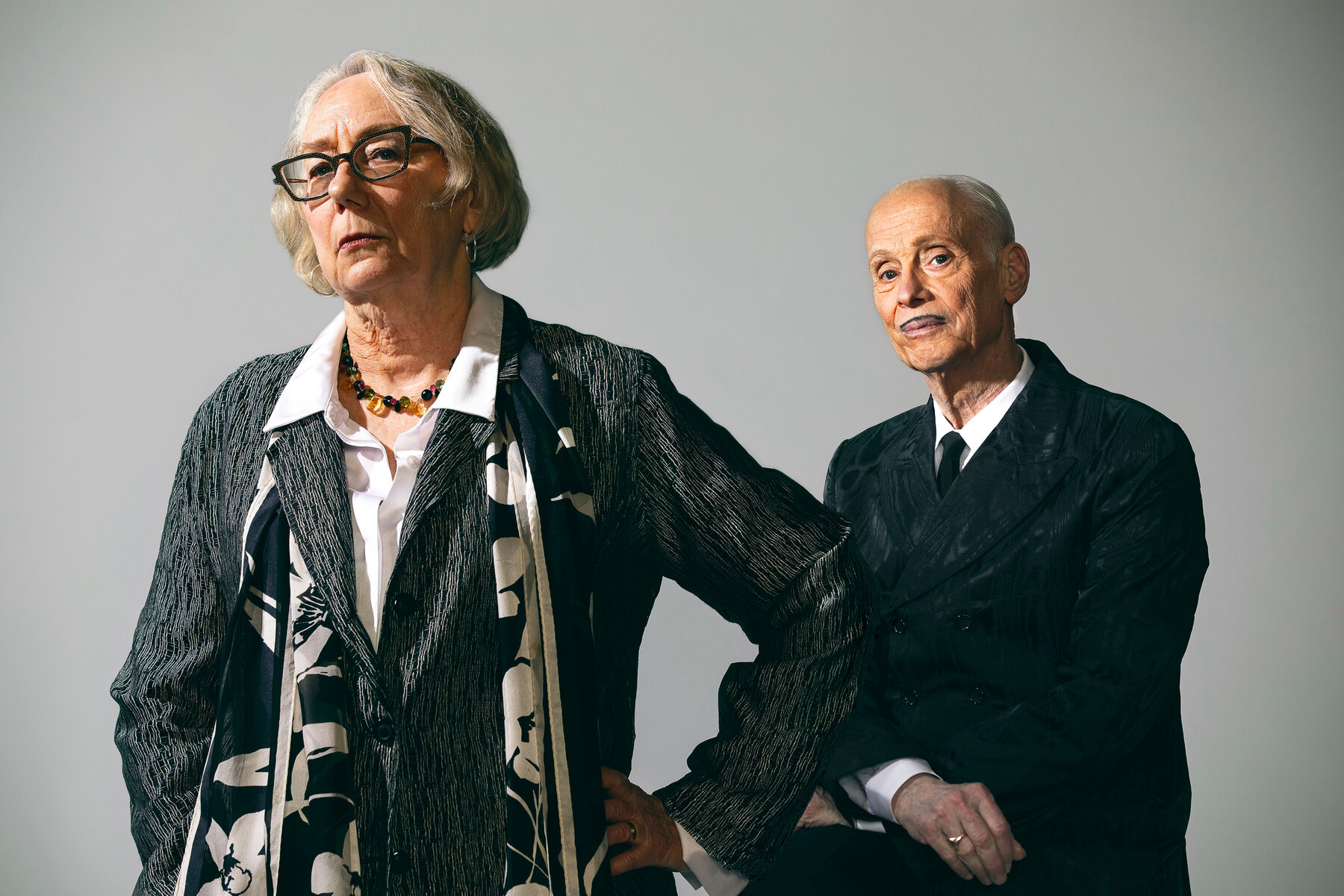 Mink Stole and John Waters, both wearing white shirts and dark gray jackets pose against a light gray background.
