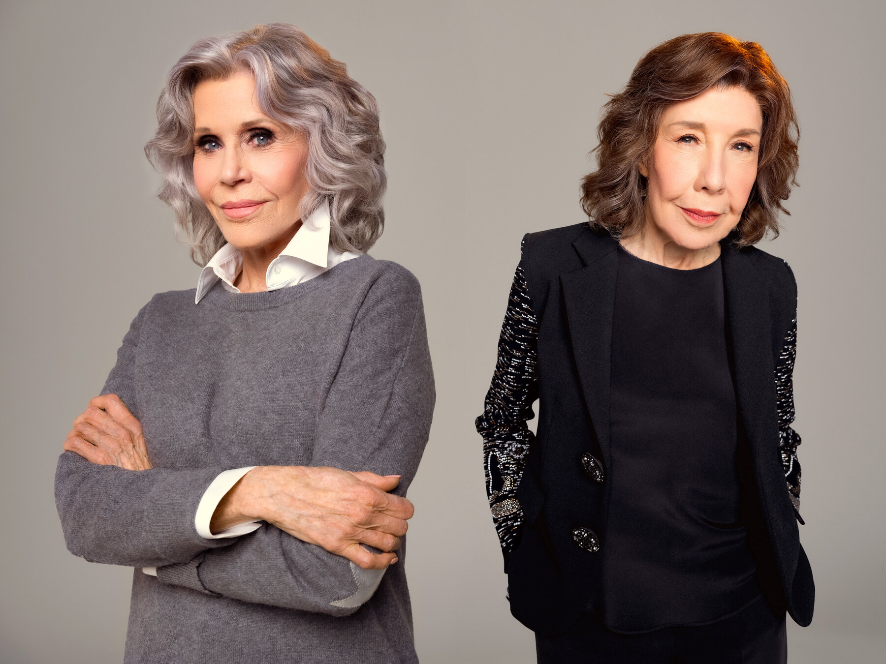 Jane Fonda and Lily Tomlin pose for a portrait. Fonda has her arms crossed and Tomlin has her hands in her pockets