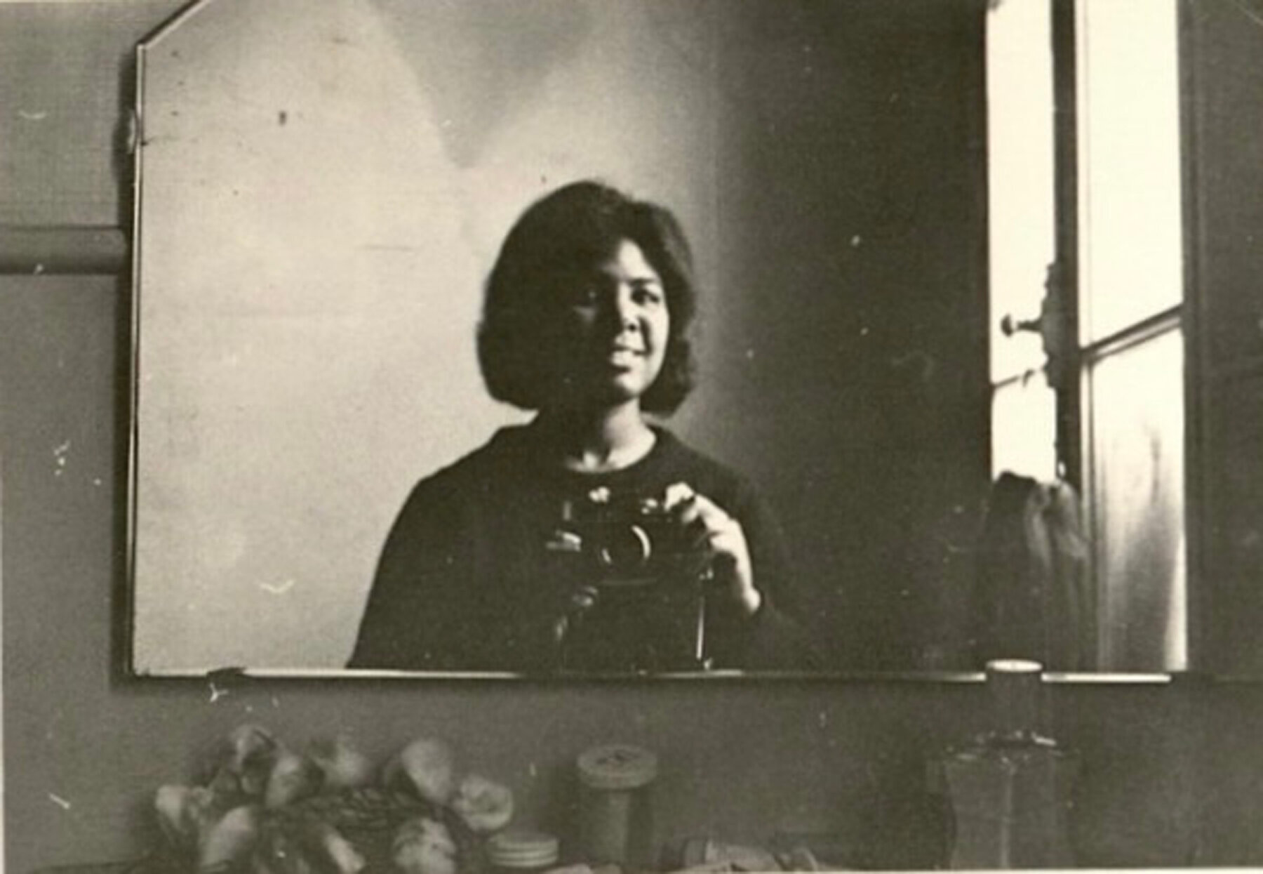 A black-and-white self-portrait of a smiling woman taken in a mirror.
