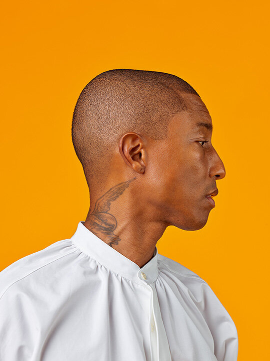 A portrait of Pharrell Williams in profile with a shaved head in front of an orange background.