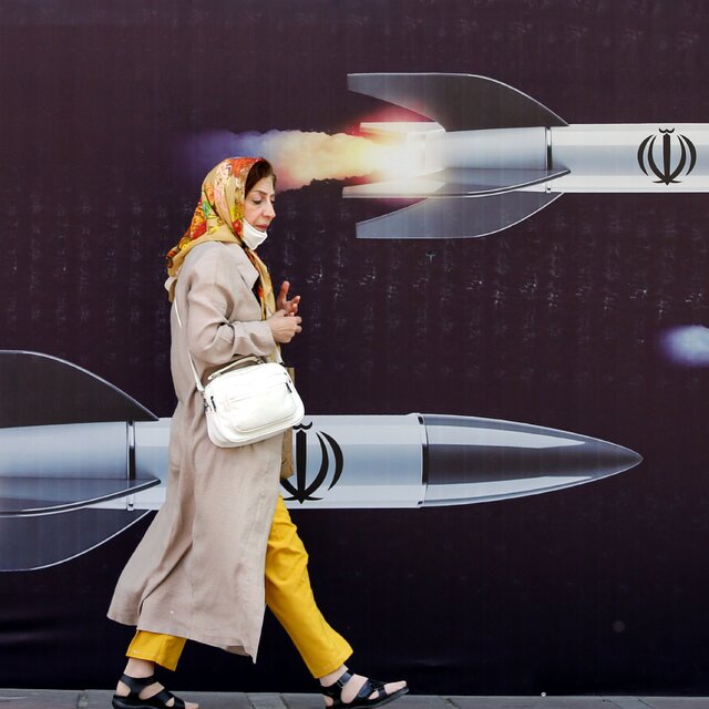 A woman in yellow pants, a tan coat, and a colorful scarf walks past a mural showing flying Iranian missiles. 