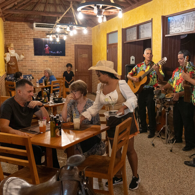 A scene at a restaurant as a woman serves two people at a table and a trio with two guitarists and a drummer play music.