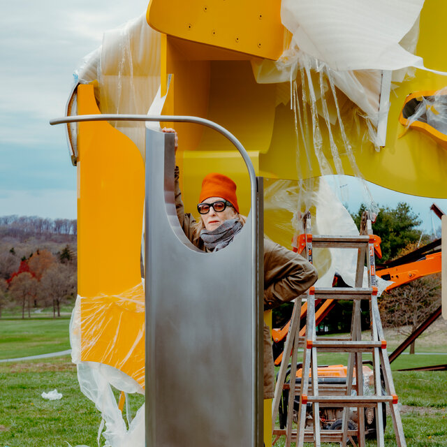 A woman in an orange cap and sunglasses stands with just her face and feet visible behind a tall piece of steel, with cutouts on top and bottom. Behind her is a ladder and next to the ladder a giant welded steel and aluminum sculpture painted two shades of yellow.