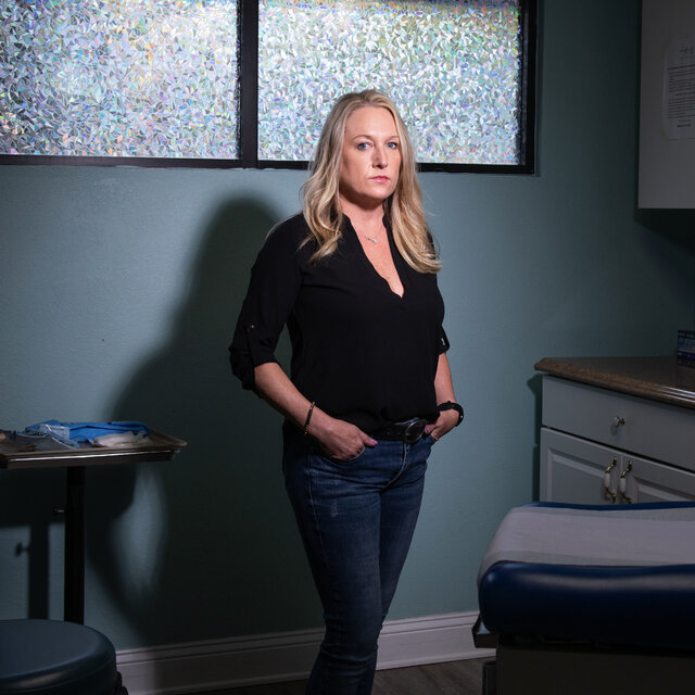 Kelly Flynn, in a black top and jeans, standing in a dimly lit medical office for a portrait.