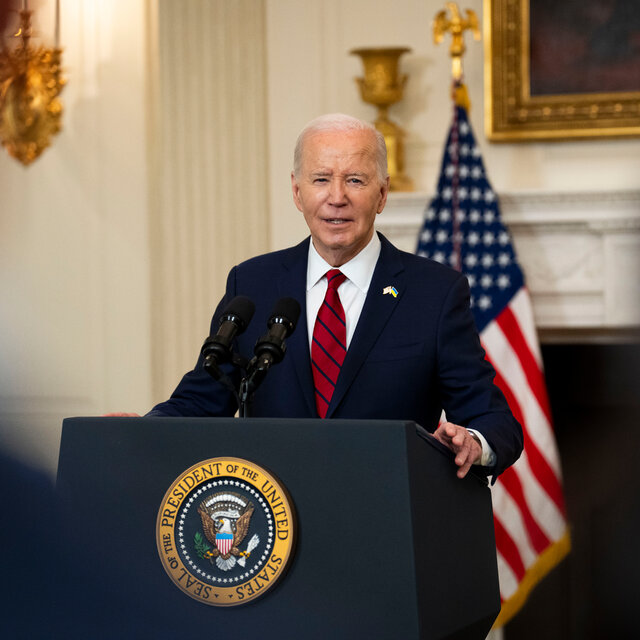 President Biden speaks at a lectern bearing the presidential seal. He is wearing a dark blue suit with a red- and blue-striped tie.