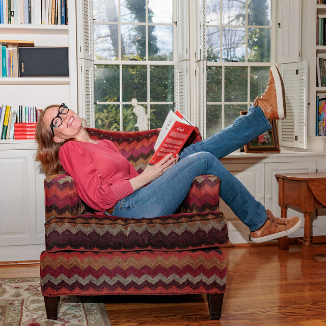 Beth Linker, wearing a bright pink blouse and jeans, lies across an armchair in her living room with one leg kicked up, holding a copy of her book.
