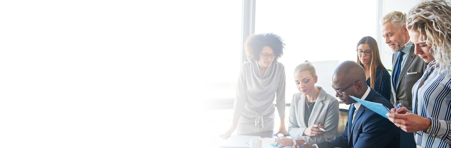 2880x1100 left white gradient of Diverse group of focused businesspeople standing around their manager sitting at an office boardroom table reviewing charts and paperwork