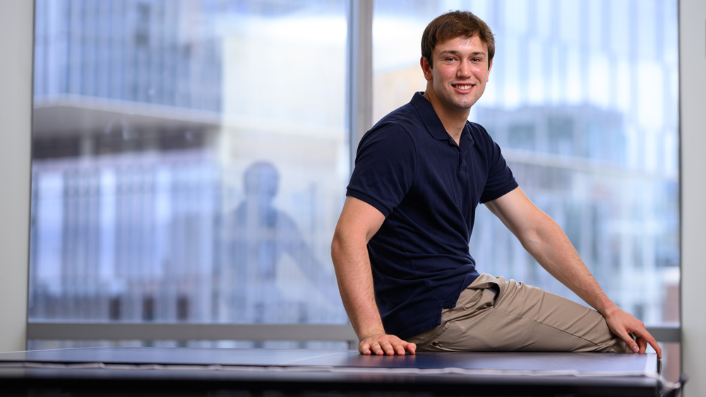 James Simon smiles while sitting on a ping pong table, with windows in background.