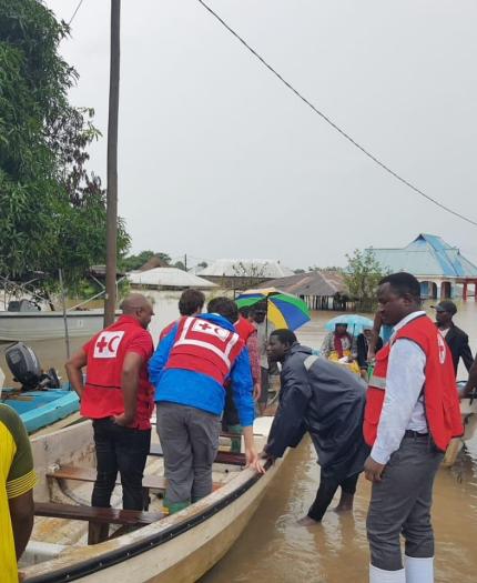 People in life vests assist others into a boat in a flooded urban area with trees and buildings visible in the background.