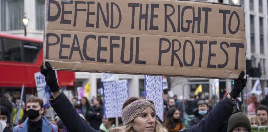 A person holding a rectangular sign that says "defend the right to peaceful protest" amongs a crowd