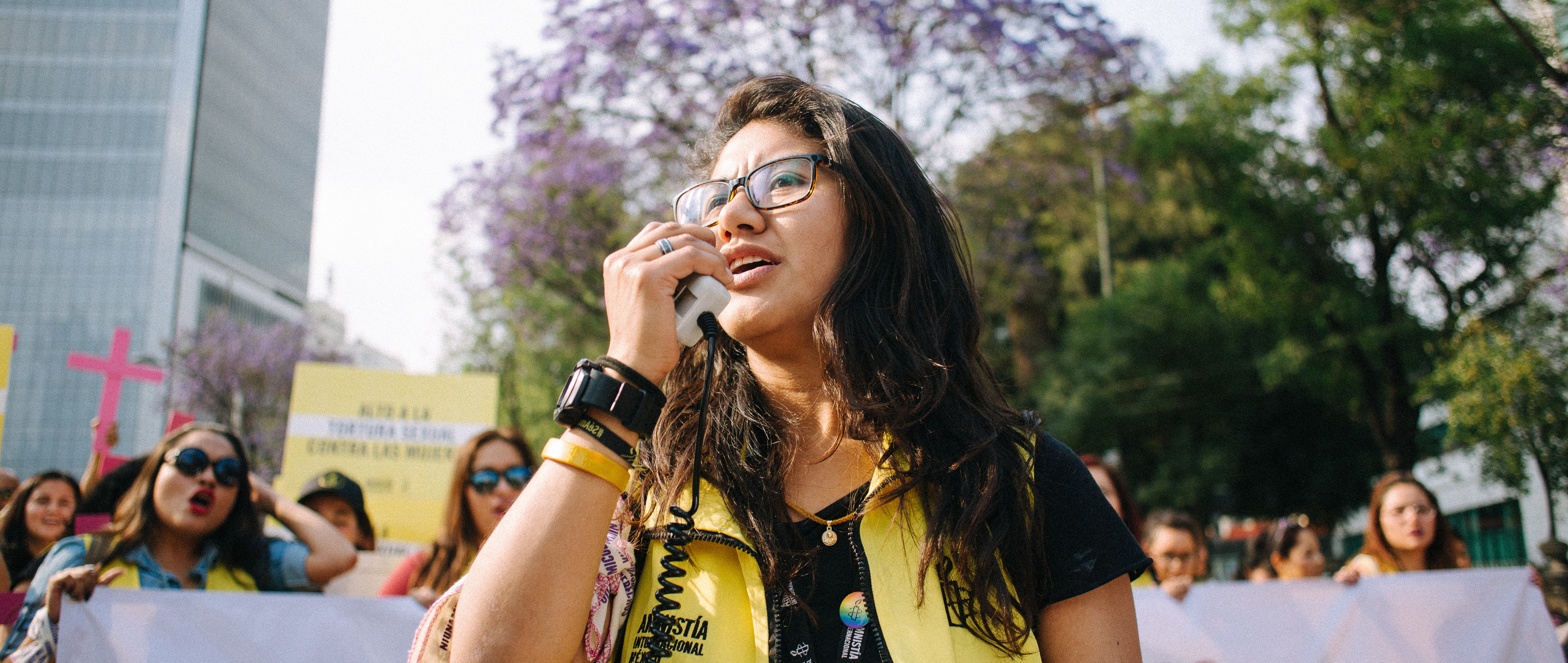 A woman speaks into a megaphone at a protest in Mexico. She has long brown hair, is wearing glasses and is wearing Amnesty branded bracelets and a yellow vest.