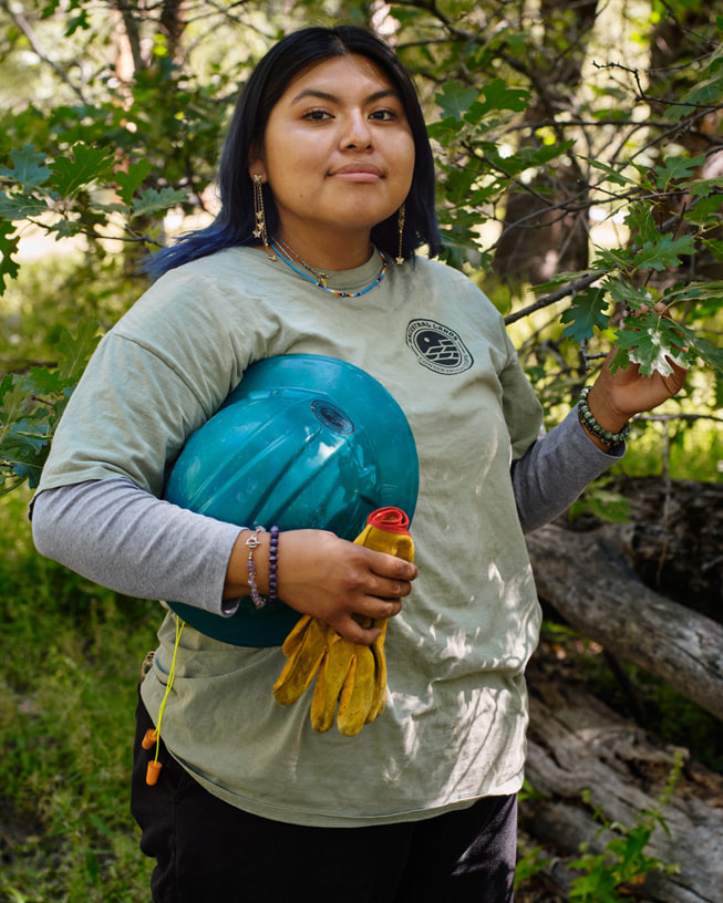 Gabrielle Draper holds her hard hat and work gloves in one hand and touches a tree branch with the other.