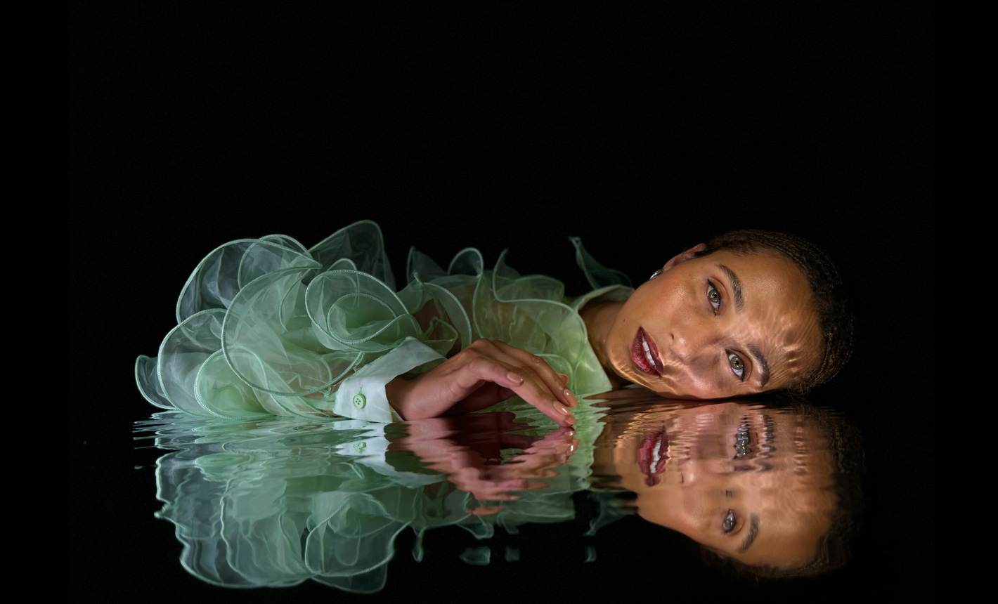 A beautifully detailed photo shot on iPhone 15 Pro of a woman laying closely to her reflection in rippled water on a black background