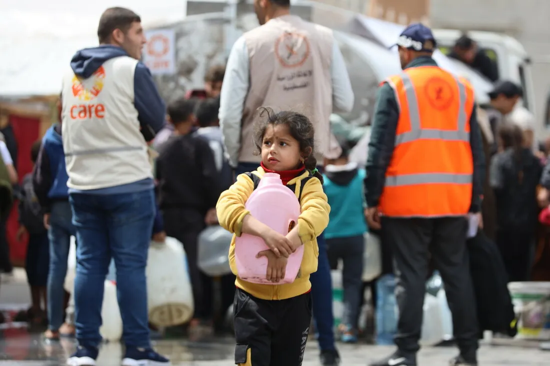 A young girl holds a pink jug close to her chest. In the background are emergency aid workers, including a man in a CARE vest.